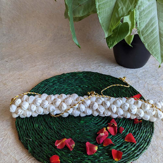 Handcrafted jasmine gajra with white flower-like detailing and golden threads, displayed on a green woven mat with scattered red rose petals.