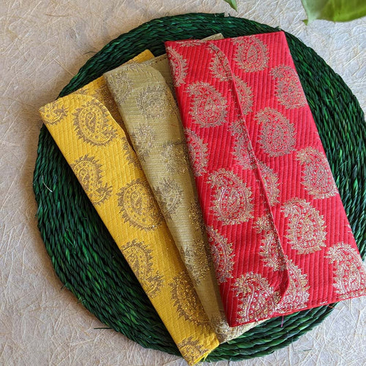 Three brocade gift pouches in red, yellow, and gold with intricate paisley patterns, displayed on a green woven mat.