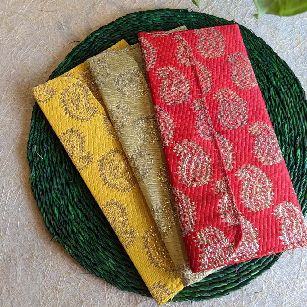 Red brocade gift pouch with gold paisley designs and zari detailing, displayed alongside yellow and gold pouches on a green woven mat.