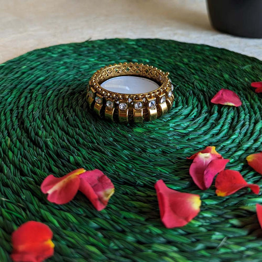 Golden Ghungroo tealight holder with crystal embellishments, placed on a green woven mat surrounded by scattered rose petals.