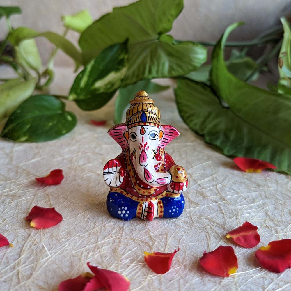 Handcrafted miniature Ganesha idol with vibrant red, blue, and gold detailing surrounded by rose petals.