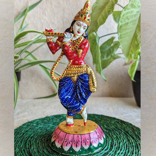 Hand-painted Krishna idol in red and blue attire, playing the flute, standing on a colorful lotus base, surrounded by green foliage.