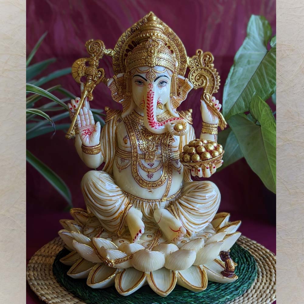Handcrafted Ganesha idol in ivory and gold finish, seated on a lotus, holding a modak and mace, displayed on a green mat with a red background and lush foliage.