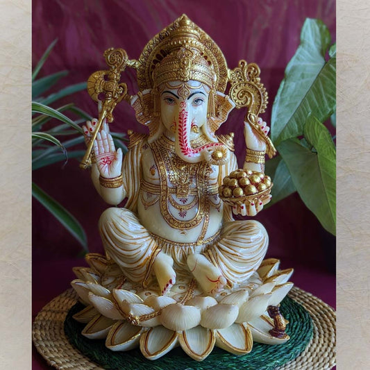 Handcrafted Ganesha idol in ivory and gold finish, seated on a lotus, holding a modak and mace, displayed on a green mat with a red background and lush foliage.