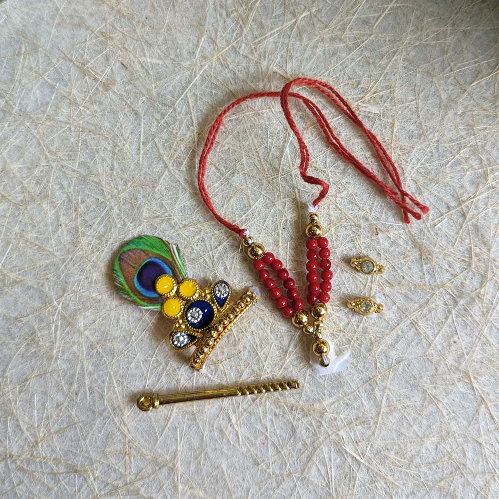 Traditional Krishna ornament set with a peacock-feather crown, red beaded necklace, flute, and decorative earrings for deity decoration.