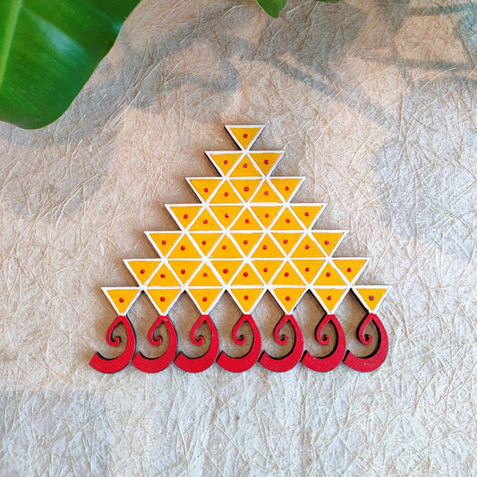 Saraswathi Kolam motif handcrafted in a triangular pattern with vibrant yellow and red detailing, placed against a textured beige background with green foliage.