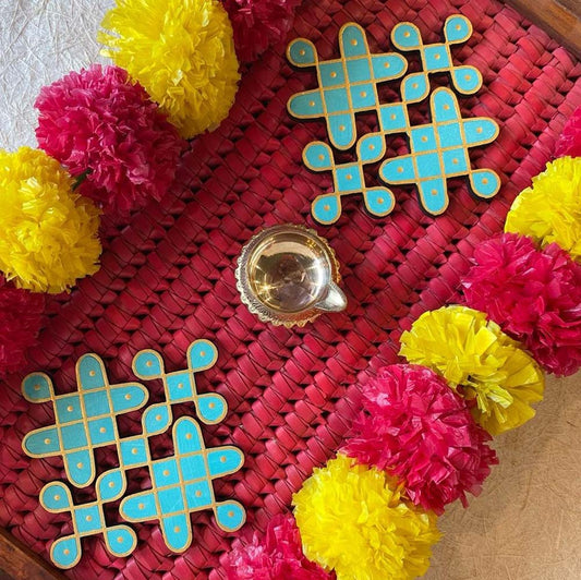 Set of two Sikku Kadal Kolam handcrafted rangoli coasters in aqua blue and gold, placed on a woven red mat with festive marigold garlands and a brass lamp.