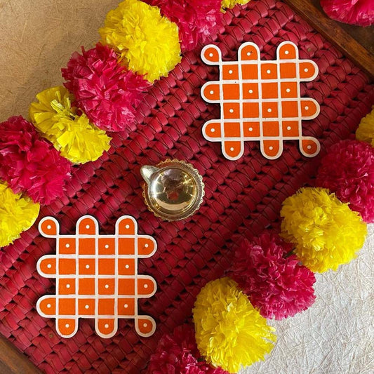 Set of two Sikku Kamala Kolam handcrafted rangoli coasters in orange and white, placed on a woven red mat with festive marigold garlands and a brass lamp.