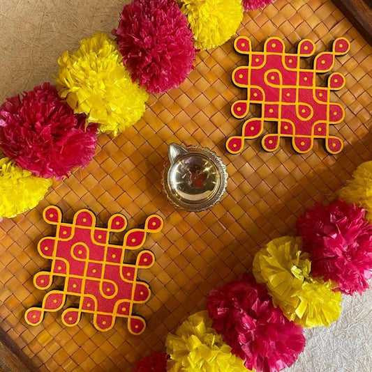 Set of two Sikku Kumkuma Kolam handcrafted rangoli coasters in red and yellow, placed on a bamboo mat with festive decorations including marigold garlands and a brass lamp.