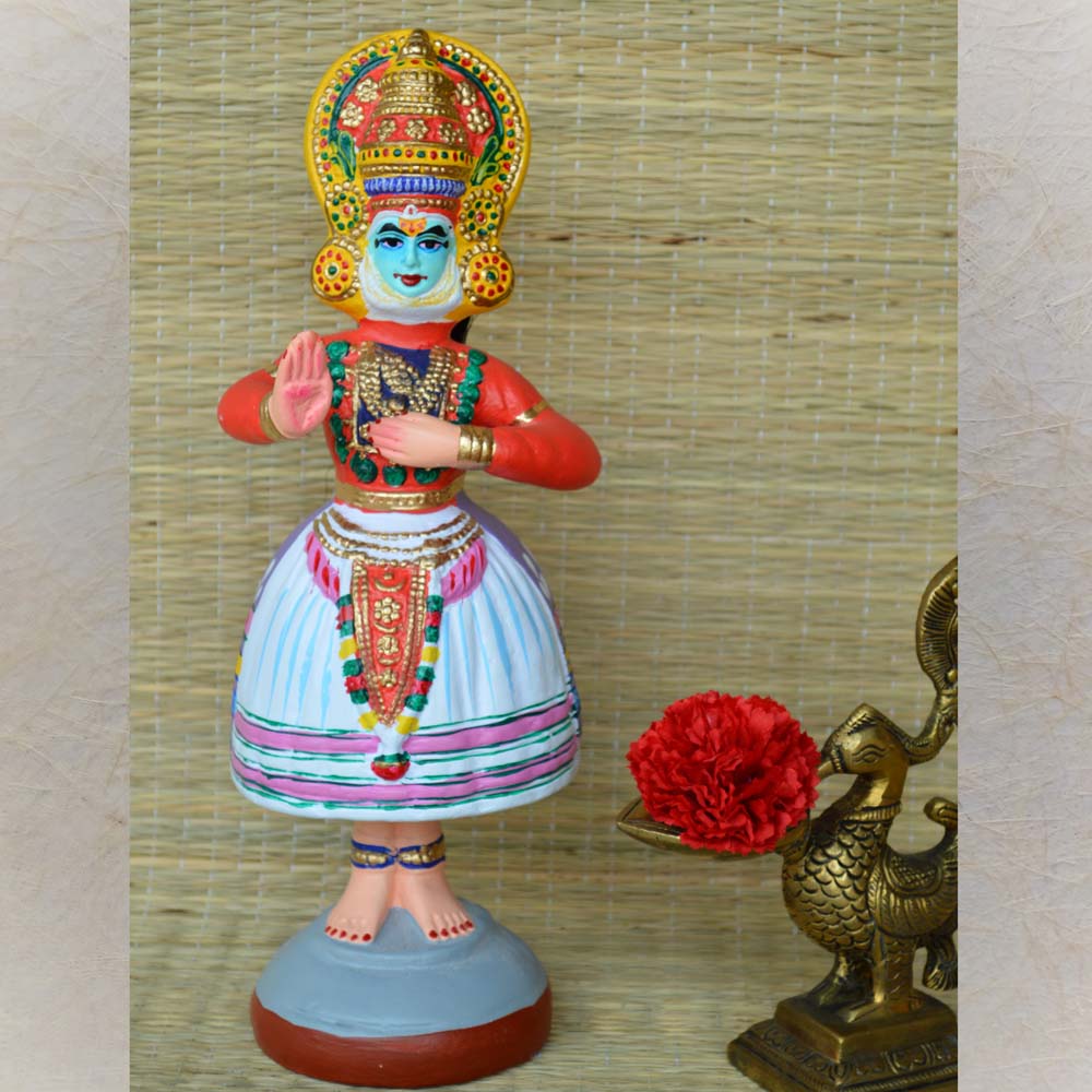 Navaratri Golu Kathakali Rama doll with an orange upper body and a white skirt adorned with colorful and golden embellishments, displayed next to a brass peacock with a red flower.