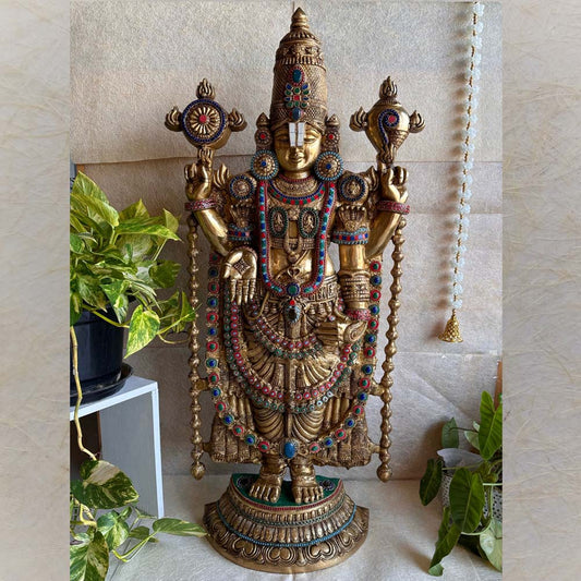 Handcrafted Balaji statue in a gold finish with colorful gemstone accents, displayed with greenery and traditional decor.
