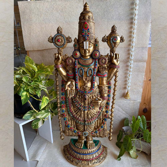 Handcrafted Balaji statue in a gold finish with colorful gemstone accents, displayed with greenery and traditional decor.