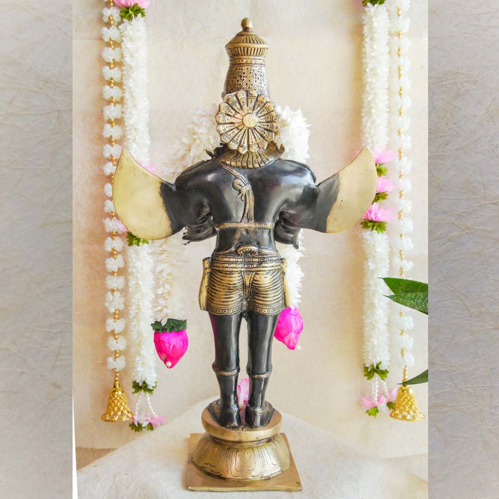 Handcrafted brass Garuda statue with intricate detailing, adorned with floral garlands and displayed with traditional decor.